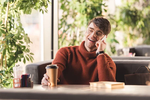 Foco seletivo de homem sorridente em óculos falando no smartphone e segurando xícara de papel com café perto do livro — Fotografia de Stock