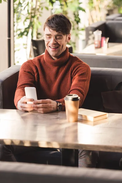 Homem sorrindo usando smartphone perto copo descartável e livro no café — Fotografia de Stock