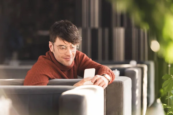 Joven sonriente en gafas usando teléfono inteligente mientras está sentado en la cafetería - foto de stock