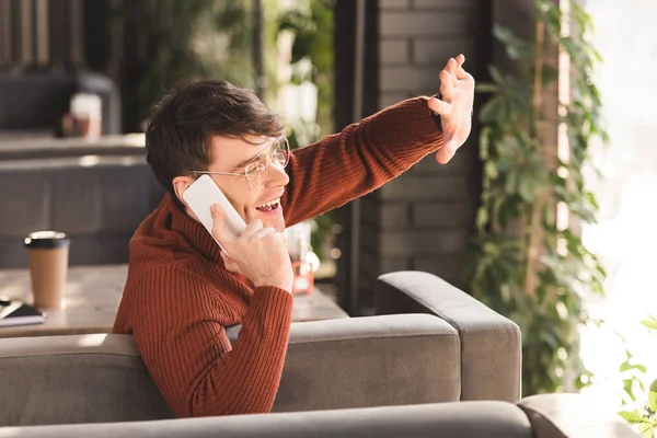 Cheerful young man in glasses talking on smartphone while waving hand in cafe — Stock Photo