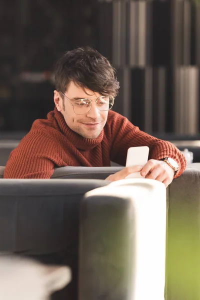 Щасливий молодий чоловік в окулярах, використовуючи смартфон, сидячи в кафе — стокове фото