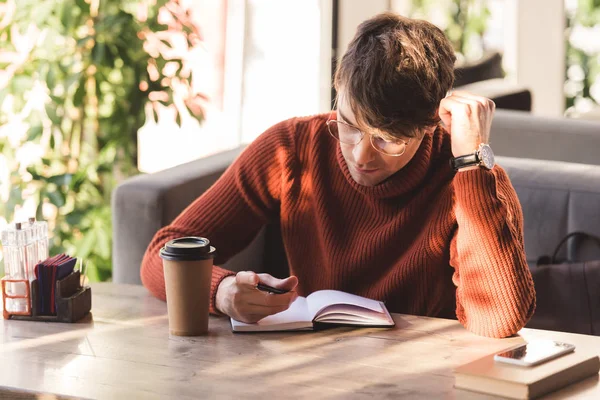 Schöner Mann mit Brille blickt in Café auf Notizbuch neben Einwegbecher — Stockfoto