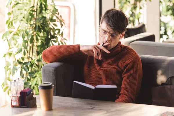 Nachdenklicher Mann mit Brille und Stift beim Blick auf Notizbuch in der Nähe von Einwegbecher im Café — Stockfoto