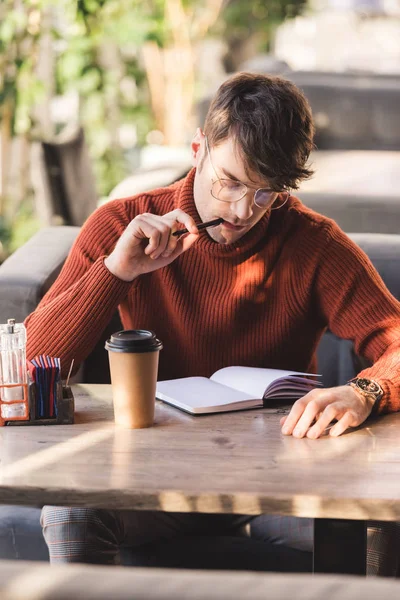 Thoughtful man in glasses holding pen near mouth while looking at notebook near disposable cup in cafe — Stock Photo