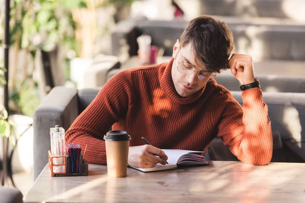 Homme réfléchi dans des lunettes écrit dans un cahier près d'une tasse jetable dans un café — Photo de stock
