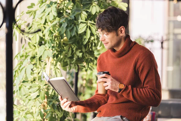 Thoughtful man in glasses looking at notebook and holding disposable cup in cafe — Stock Photo