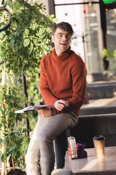 Щасливий молодий чоловік в окулярах тримає блокнот, стоячи біля одноразової чашки в кафе — стокове фото