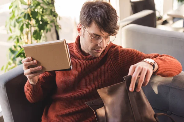 Homem bonito em óculos segurando livros e olhando para o saco no café — Fotografia de Stock