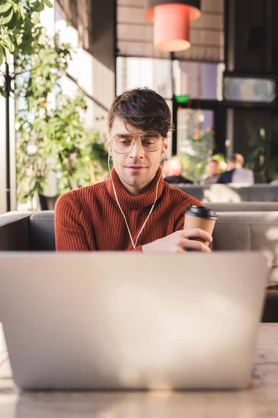 Foyer sélectif de l'homme écouter de la musique dans les écouteurs tout en utilisant un ordinateur portable et tenant tasse en papier dans le café — Photo de stock