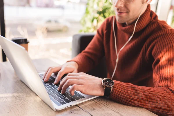 Vista recortada del hombre escuchando música en los auriculares mientras usa el ordenador portátil en la cafetería - foto de stock