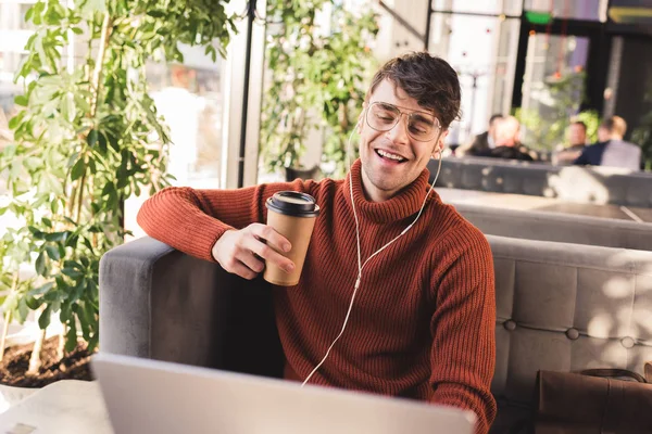 Hombre sonriente escuchando música en los auriculares mientras usa el ordenador portátil y sostiene la taza desechable en la cafetería - foto de stock