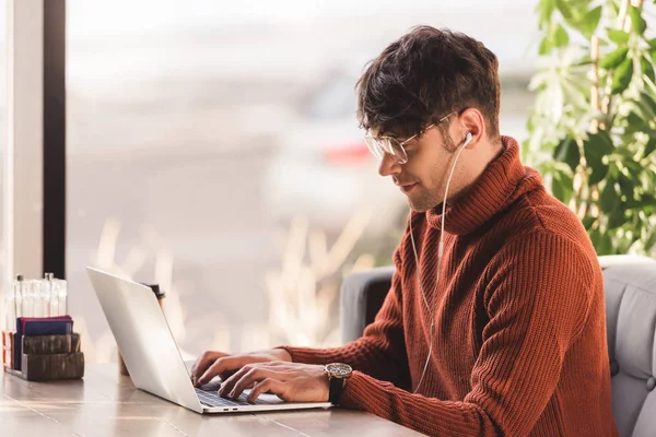 Jovem feliz ouvindo música em fones de ouvido e usando laptop no café — Fotografia de Stock