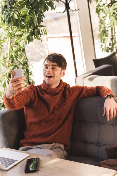 Homme gai dans des lunettes prenant selfie près d'un ordinateur portable dans un café — Photo de stock