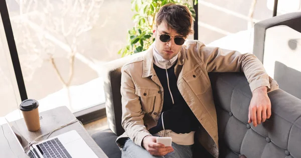 Hombre serio en gafas de sol sosteniendo teléfono inteligente mientras está sentado en la cafetería - foto de stock