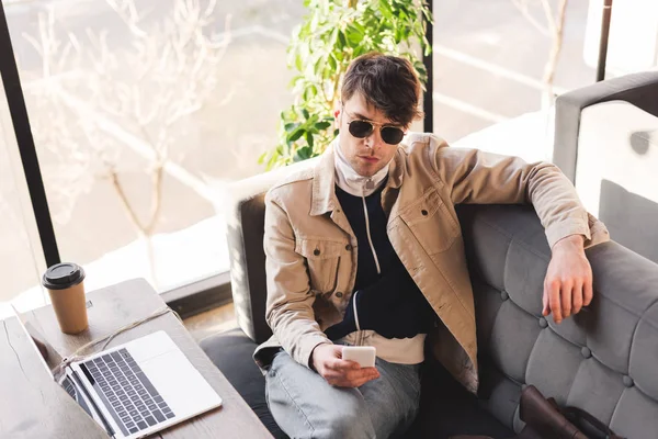 Homme élégant dans des lunettes de soleil tenant smartphone tout en étant assis dans le café — Photo de stock