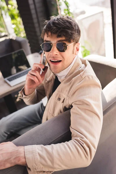 Вибірковий фокус людини в сонцезахисних окулярах, що розмовляють на смартфоні біля ноутбука в кафе — стокове фото