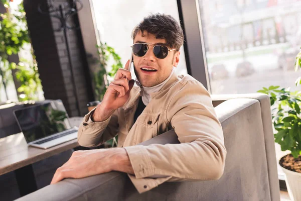 Selective focus of trendy man in sunglasses talking on smartphone near laptop in cafe — Stock Photo