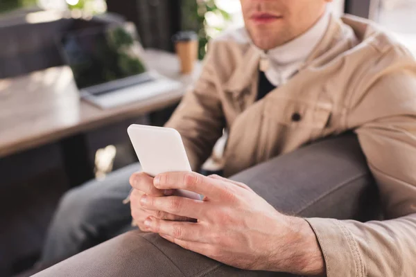 Cropped view of trendy man sitting near laptop and paper cup and using smartphone — Stock Photo