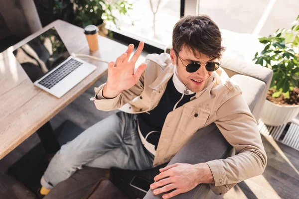 Enfoque selectivo del hombre de moda en gafas de sol sentado cerca de la computadora portátil y taza de papel mientras agita la mano en la cafetería - foto de stock