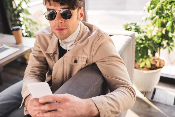 Вибірковий фокус модного чоловіка в сонцезахисних окулярах за допомогою смартфона в кафе — стокове фото