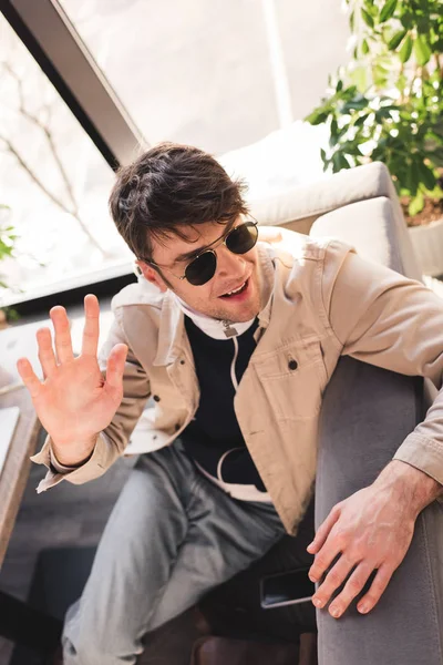 Selective focus of trendy man in sunglasses sitting near smartphone with blank screen and waving hand in cafe — Stock Photo