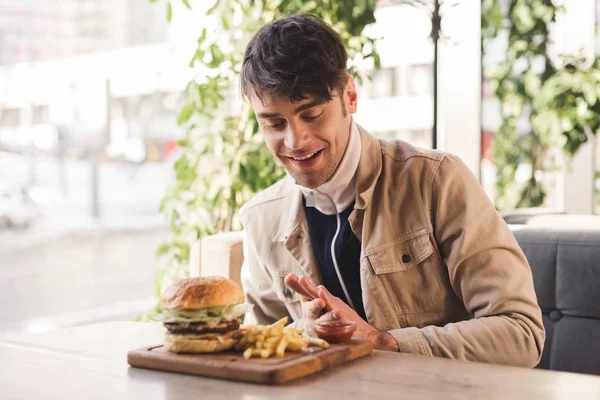Homme heureux regardant des frites près de délicieux hamburger sur la planche à découper dans le café — Photo de stock
