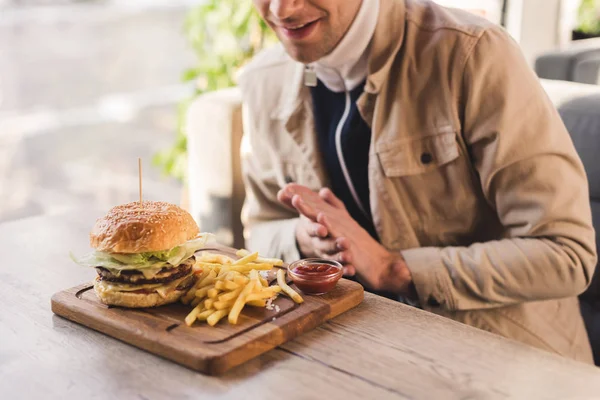 Vista recortada de hombre alegre mirando sabrosa hamburguesa y papas fritas en la tabla de cortar en la cafetería - foto de stock