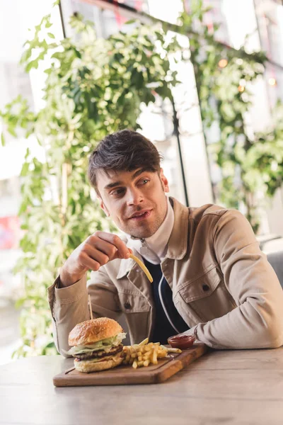 Joven guapo sosteniendo freír francés cerca de sabrosa hamburguesa en la tabla de cortar en la cafetería - foto de stock