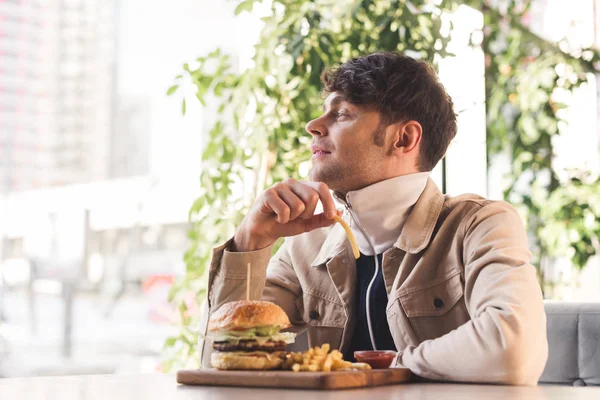 Joyeux jeune homme tenant des frites près de hamburger savoureux sur la planche à découper dans le café — Photo de stock