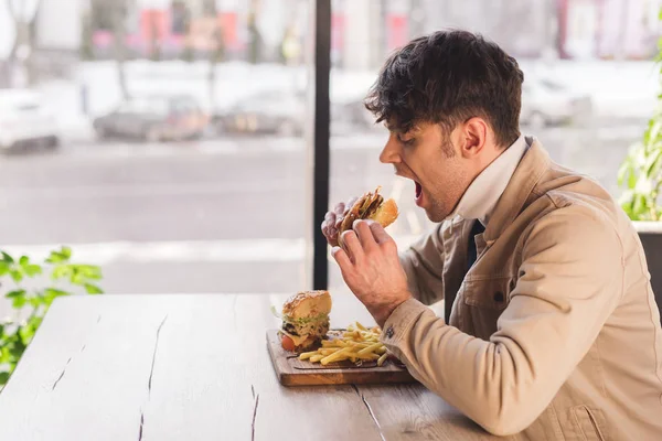 Handsome man eating tasty burger in cafe — Stock Photo