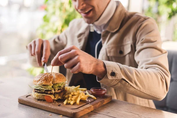 Cropped view of cheerful young man cutting tasty burger on cutting board in cafe — Stock Photo