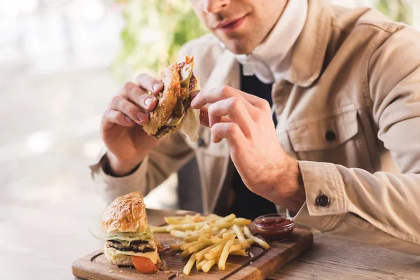 Vista recortada de un joven sosteniendo sabrosa hamburguesa cerca de papas fritas en la tabla de cortar en la cafetería - foto de stock