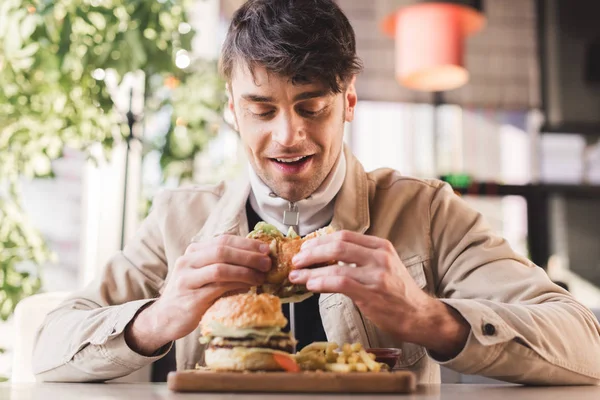 Messa a fuoco selettiva di felice giovane guardando gustoso hamburger vicino patatine fritte sul tagliere in caffè — Foto stock