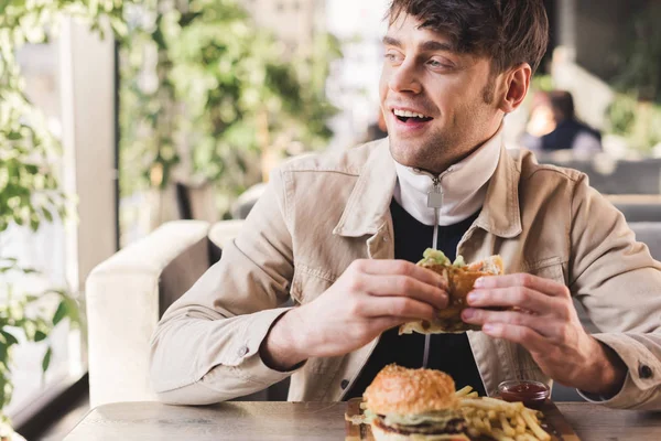 Selective focus of happy young man holding tasty burger near french fries on cutting board in cafe — Stock Photo