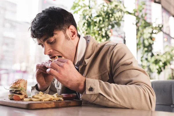 Junger Mann isst leckeren Hamburger in der Nähe von Pommes auf Schneidebrett in Café — Stockfoto