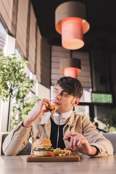 Selective focus of handsome young man holding tasty hamburger near french fries on cutting board in cafe — Stock Photo