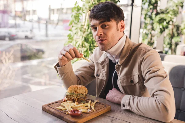 Allegro uomo che tiene friggere francese vicino delizioso hamburger sul tagliere in caffè — Foto stock