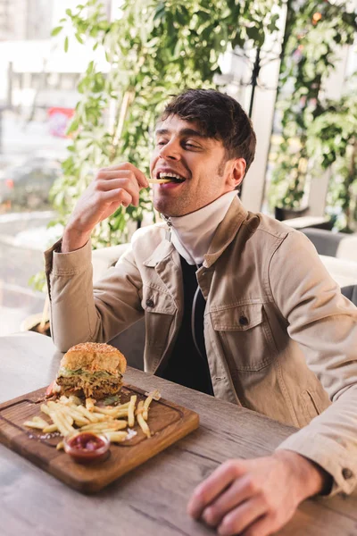 Homem feliz comer batata frita perto delicioso hambúrguer na placa de corte no café — Fotografia de Stock