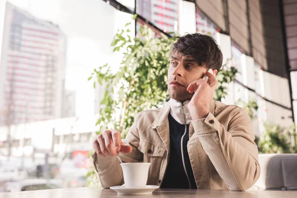 Мужчина держит ложку возле чашки с капучино и разговаривает на смартфоне в кафе — стоковое фото