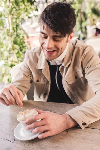 Hombre alegre sosteniendo cuchara en taza con capuchino en la cafetería - foto de stock