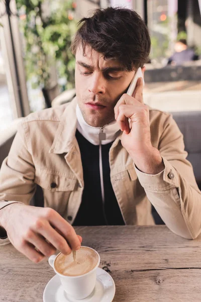 Hombre serio sosteniendo cuchara en taza con capuchino y hablando en el teléfono inteligente en la cafetería - foto de stock