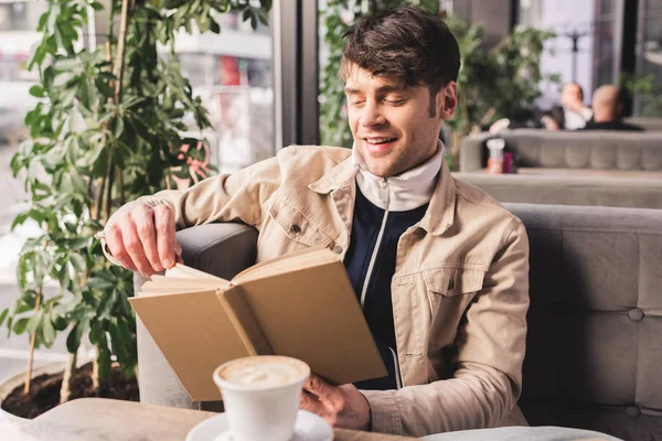 Hombre feliz sosteniendo libro cerca de la taza con capuchino en la cafetería - foto de stock