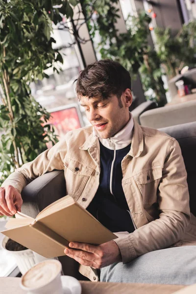 Glücklicher Mann liest Buch neben Tasse mit Cappuccino im Café — Stockfoto