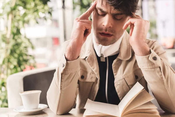 Hombre enfocado leer libro cerca de la taza con capuchino en la cafetería - foto de stock