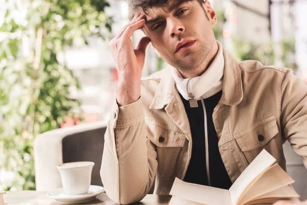 Homem pensativo sentado perto de livro e xícara com cappuccino no café — Fotografia de Stock