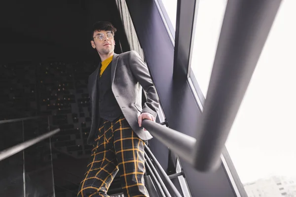 Joven hombre con estilo en gafas de pie en las escaleras y mirando a la ventana - foto de stock