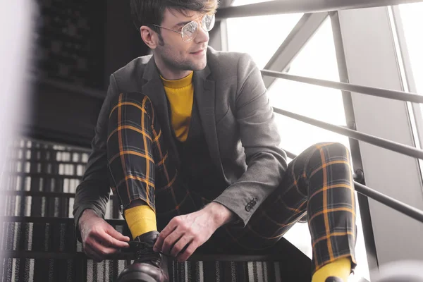 Щасливий стильний чоловік в окулярах сидить на сходах і торкається шнурівки, дивлячись вікна — стокове фото
