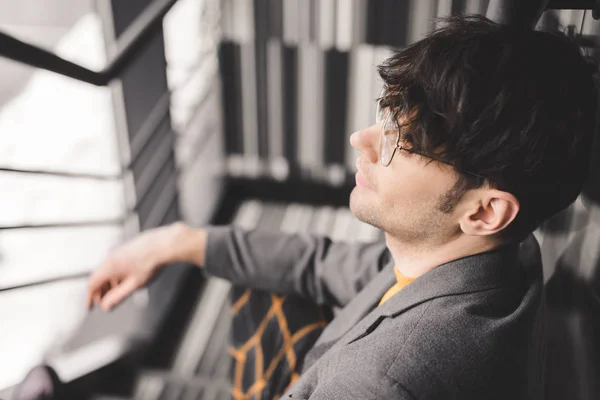 Selective focus of dreamy young man in glasses sitting on stairs — Stock Photo