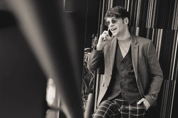Трогательный мужчина в солнцезащитных очках улыбается, стоя на лестнице и разговаривая по смартфону, черно-белая фотография — стоковое фото