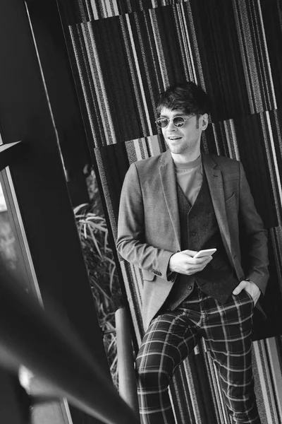 Trendy man in sunglasses standing with hand in pocket on stairs and holding smartphone, black and white photography — Stock Photo